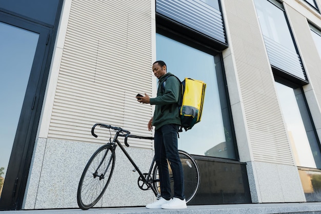 Un mensajero afroamericano con una mochila amarilla camina con una bicicleta a lo largo de un edificio de oficinas