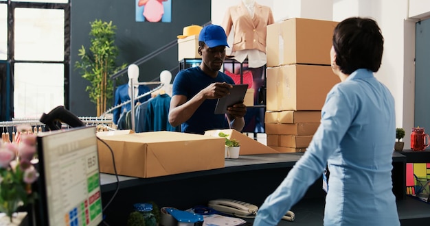 Mensajero afroamericano tomando fotografías de cajas de cartón usando una tableta antes de poner al gerente de la tienda a firmar el informe de entrega. Empleado preparando paquetes para envío en boutique moderna