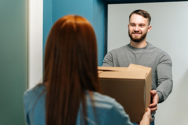 Mensajero barbudo sonriente que entrega un paquete de caja de cartón a una clienta irreconocible en la puerta de su casa