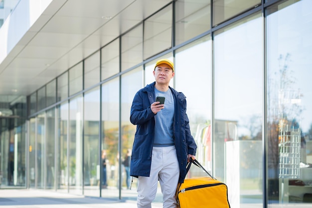 Mensajero de comida masculino asiático que usa el móvil usando la aplicación de teléfono móvil en el teléfono inteligente mientras está de pie en la calle de la ciudad con aislamiento con mochila térmica entregando alimentos. buscando la dirección del cliente. comprar mapa en línea