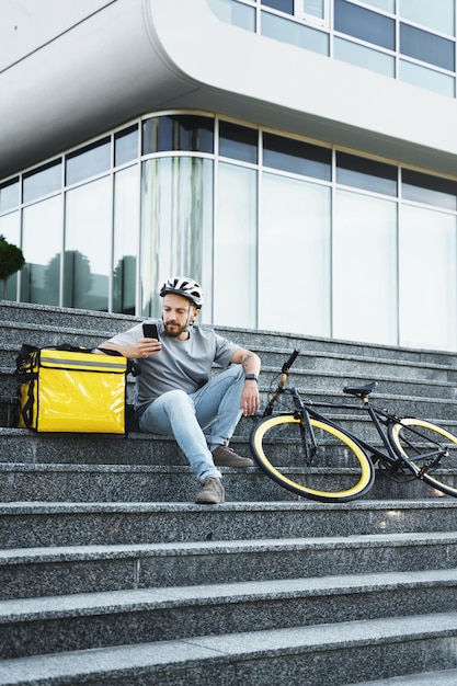 Mensajero de entrega de alimentos exprés sentado en las escaleras con bolsa aislada y bicicleta