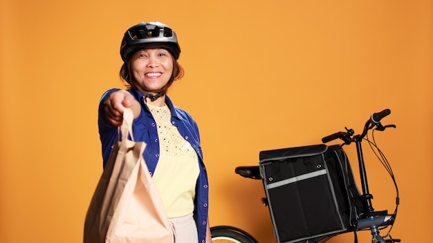 El mensajero espera frente a la puerta del apartamento para que el cliente recoja el pedido de comida para llevar. El ciclista de entrega de comida ofrece una bolsa de almuerzo para llevar al cliente, aislado sobre el fondo de un estudio naranja