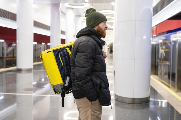 El mensajero está esperando el metro. Un hombre con barba y una mochila amarilla entrega comida por la ciudad.