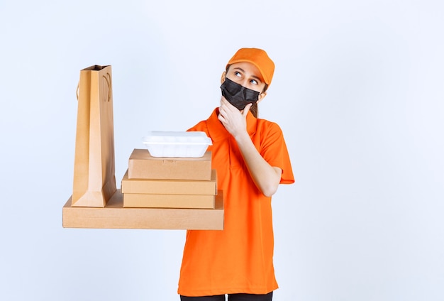 Mensajero femenino en uniforme amarillo y máscara negra sosteniendo varios paquetes de cartón, caja de comida para llevar y bolsa de compras mientras se ve confundido y pensativo