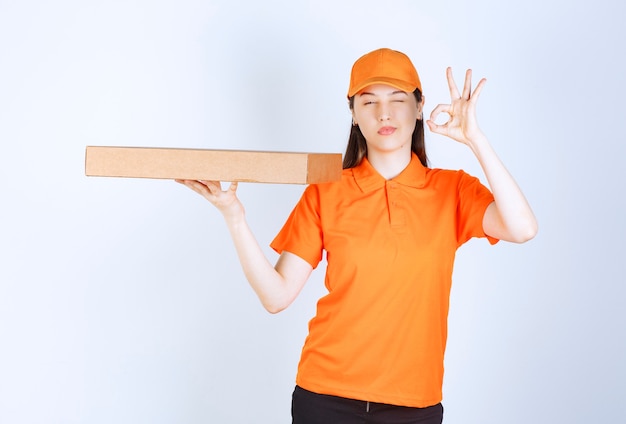 Mensajero femenino en uniforme amarillo sosteniendo una caja de cartón para llevar y mostrando el signo de la mano de disfrute.