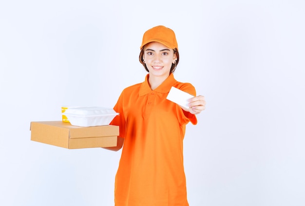 Mensajero femenino en uniforme naranja sosteniendo cajas de comida para llevar amarillas y blancas, paquete de cartón y presentando su tarjeta de visita