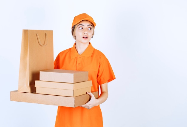 Mensajero femenino en uniforme naranja sosteniendo un stock de paquetes de cartón y bolsas de compras y parece confundido y aterrorizado