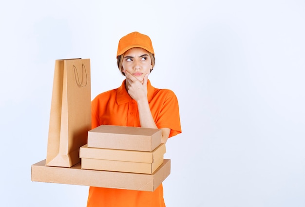 Mensajero femenino en uniforme naranja sosteniendo un stock de paquetes de cartón y bolsas de compras y parece confundido y aterrorizado