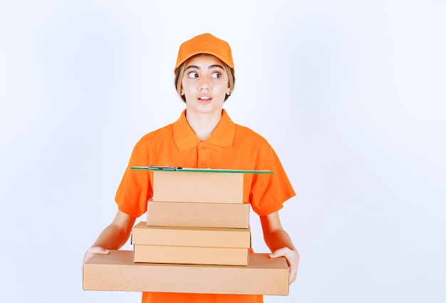 Mensajero femenino en uniforme naranja sosteniendo un stock de paquetes de cartón y parece confundido y pensativo