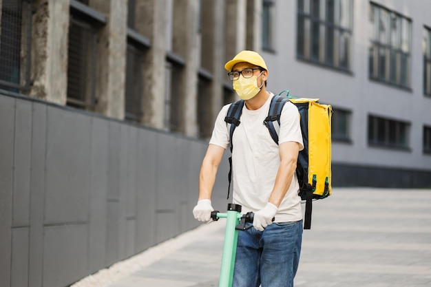 Mensajero de hombre con mascarilla amarilla en scooter en las calles de la ciudad con una entrega de comida caliente