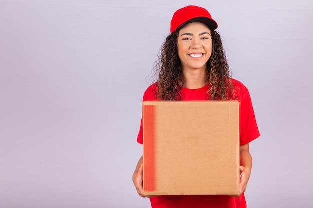 Mensajero joven hermosa que entrega un paquete enorme. Una mujer de entrega con un uniforme rojo está sonriendo con una gran caja de cartón en sus manos. Las compras en línea. Igualdad de mujeres.