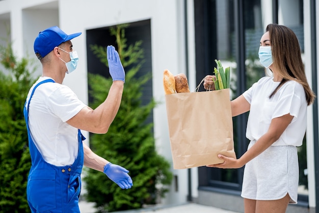 Mensajero joven con máscara protectora y guantes entrega mercancías a una mujer joven durante la cuarentena