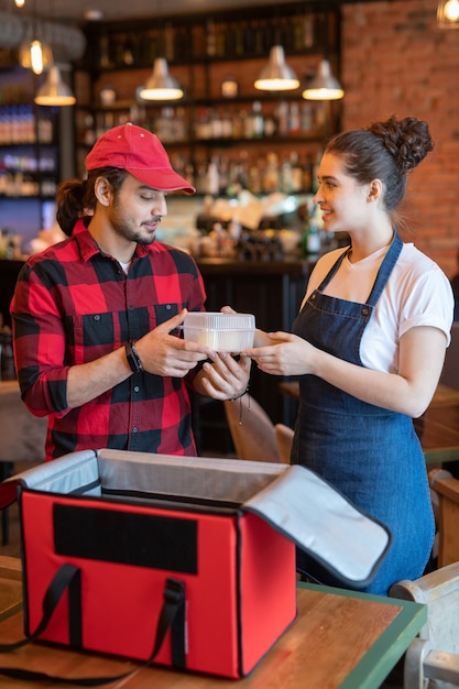 Mensajero joven en ropa de trabajo teniendo recipiente de plástico con comida de manos de camarera ayudándolo a empacar pedidos de clientes en café