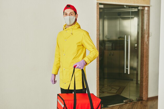 Mensajero joven en uniforme con gran bolsa roja con pedidos de clientes mientras está de pie junto a la pared de la oficina moderna y entregando almuerzos