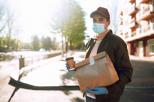 El mensajero con máscara protectora y guantes médicos en un scooter entrega un paquete de papel artesanal con comida. Servicio de entrega bajo cuarentena, brote de enfermedad, condiciones de pandemia de coronavirus covid-19.