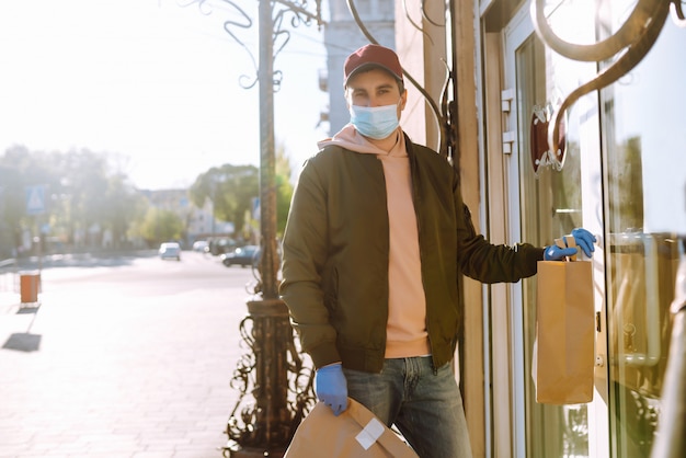 El mensajero con máscara protectora y guantes médicos en un scooter entrega un paquete de papel artesanal con comida. Servicio de entrega bajo cuarentena, brote de enfermedad, condiciones de pandemia de coronavirus covid-19.