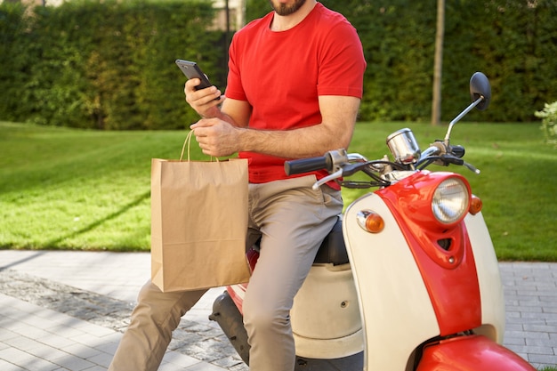 Mensajero masculino mediante aplicación móvil mientras entrega ingenio de bolsa ecológica de papel