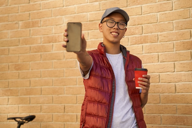 Mensajero masculino asiático alegre joven con la taza de café que muestra scr
