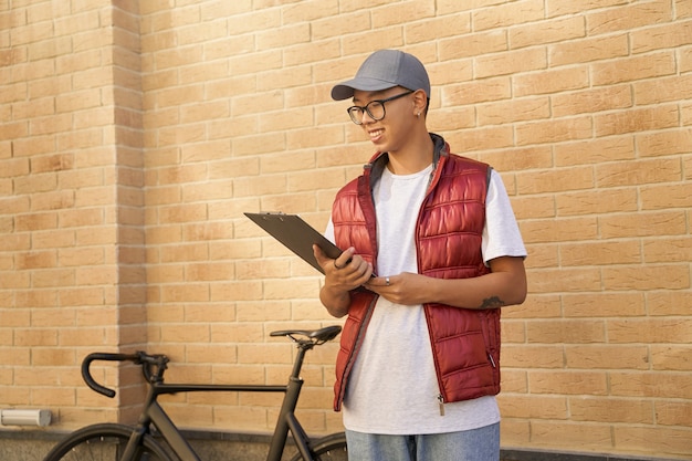 Mensajero masculino asiático joven que mira el portapapeles mientras está de pie aga