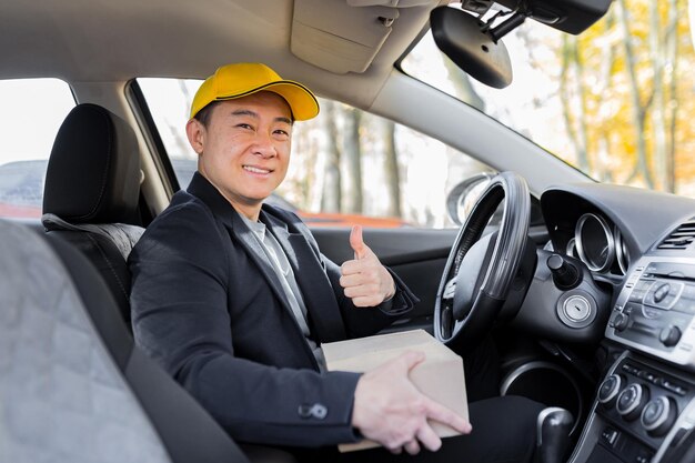 El mensajero masculino asiático mira a la cámara y sonríe sosteniendo un paquete sentado en el auto