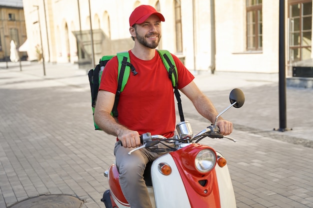 Mensajero masculino caucásico alegre joven con bolsa termo entregando comida a los clientes en moto, cabalgando por la calle soleada de la ciudad y sonriendo. Concepto de servicios de entrega