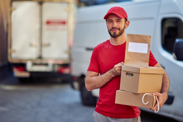 Mensajero masculino caucásico guapo joven en uniforme rojo con paquetes y bolsa de papel para la entrega