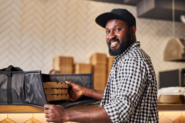 Mensajero masculino guapo mirando a la cámara y sonriendo mientras empaca cajas de pizza de cartón en una bolsa térmica aislada