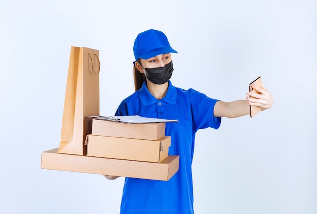 Mensajero mujer con máscara y uniforme azul sosteniendo una bolsa de cartón y varias cajas mientras toma su selfie.