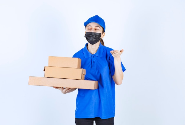 Mensajero mujer con máscara y uniforme azul sosteniendo un stock de cajas de cartón y oliendo el producto.