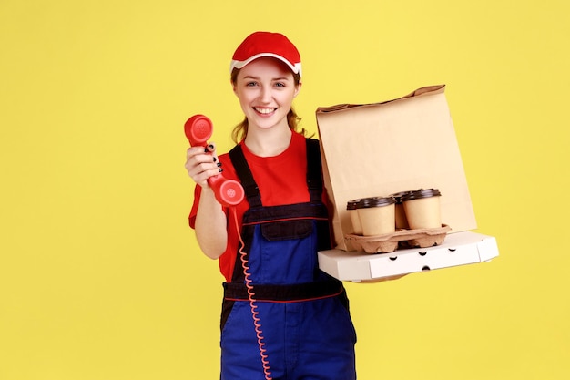 Mensajero de mujer sosteniendo teléfono fijo y café con llamada de pizza para pedir comida rápida mirando a la cámara con una sonrisa y expresión feliz Disparo de estudio interior aislado sobre fondo amarillo