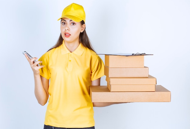 Mensajero mujer en uniforme amarillo con montón de cajas de cartón y teléfono móvil.