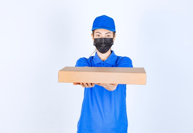 Mensajero mujer en uniforme azul y mascarilla entregando una caja de cartón al cliente.