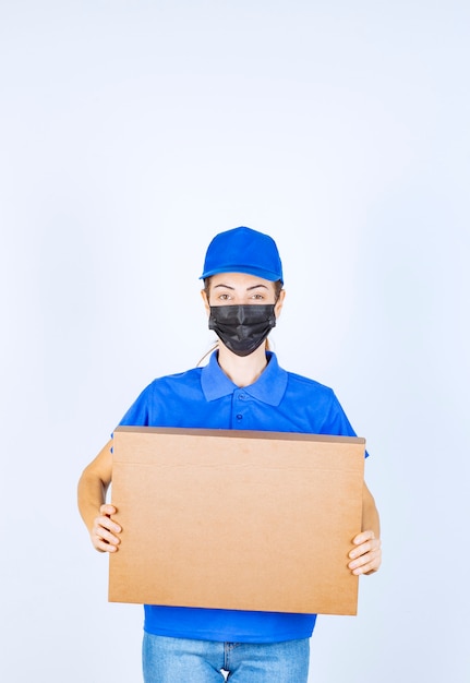 Mensajero mujer en uniforme azul y mascarilla entregando una caja de cartón al cliente.