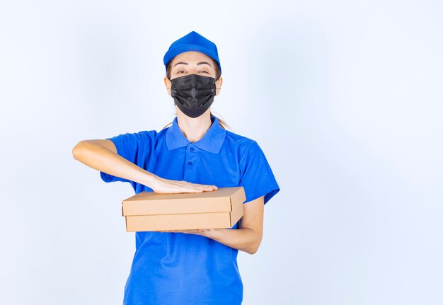 Mensajero mujer en uniforme azul y mascarilla entregando una caja de cartón al cliente.