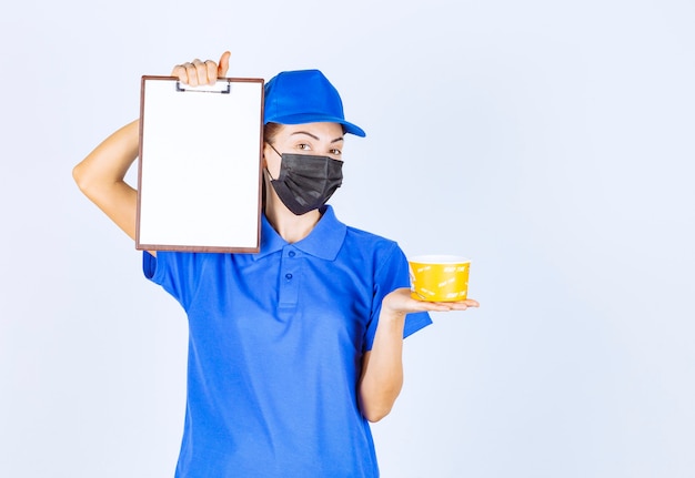 Mensajero mujer en uniforme azul y mascarilla entregando comida para llevar y pidiendo al cliente que firme.