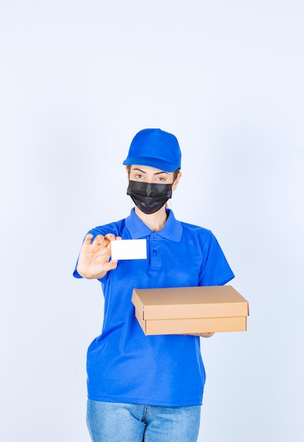 Mensajero mujer en uniforme azul y mascarilla entregando un paquete de cartón y presentando su tarjeta de visita.