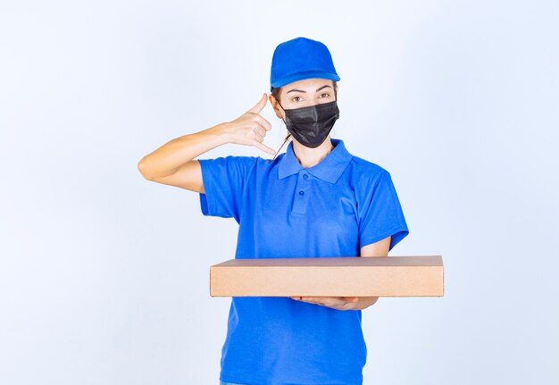 Mensajero mujer en uniforme azul y mascarilla sosteniendo una caja de cartón y pidiendo una llamada.