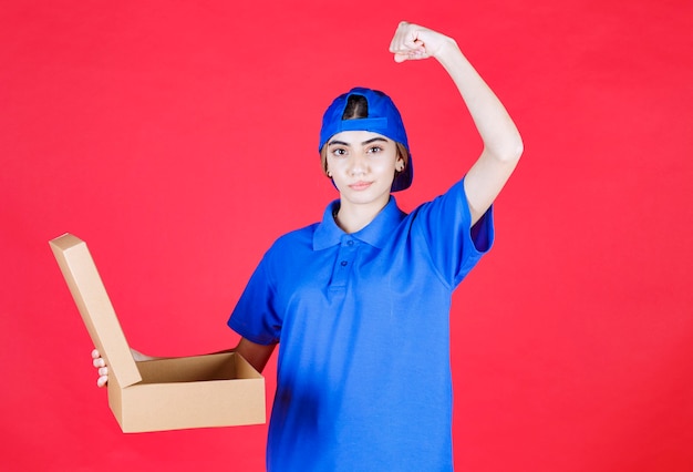 Mensajero mujer en uniforme azul sosteniendo una caja de cartón para llevar y mostrando su músculo del brazo.