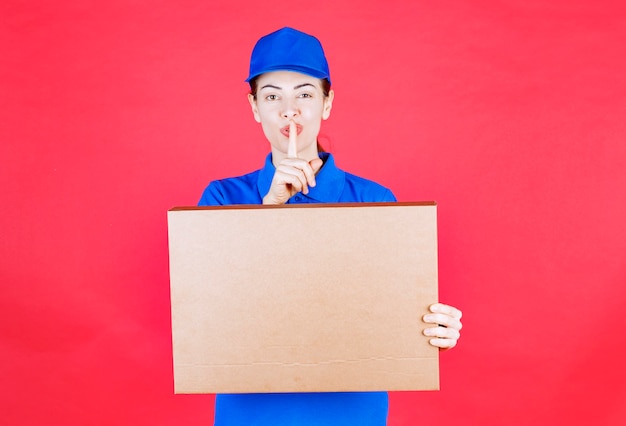 Mensajero mujer en uniforme azul sosteniendo una caja de cartón de pizza para llevar y pidiendo silencio.