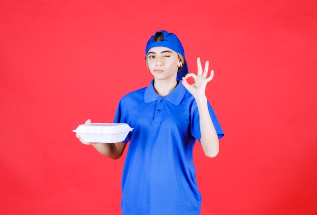 Mensajero mujer en uniforme azul sosteniendo una caja de comida para llevar blanca y mostrando signo de mano de disfrute.