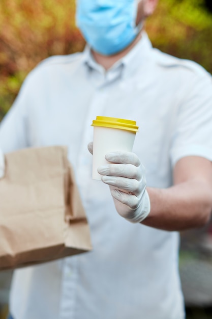 El mensajero, repartidor con máscara protectora y guantes médicos entrega comida para llevar y café. Servicio de entrega bajo cuarentena, brote de enfermedad, condiciones de pandemia de coronavirus covid-19.