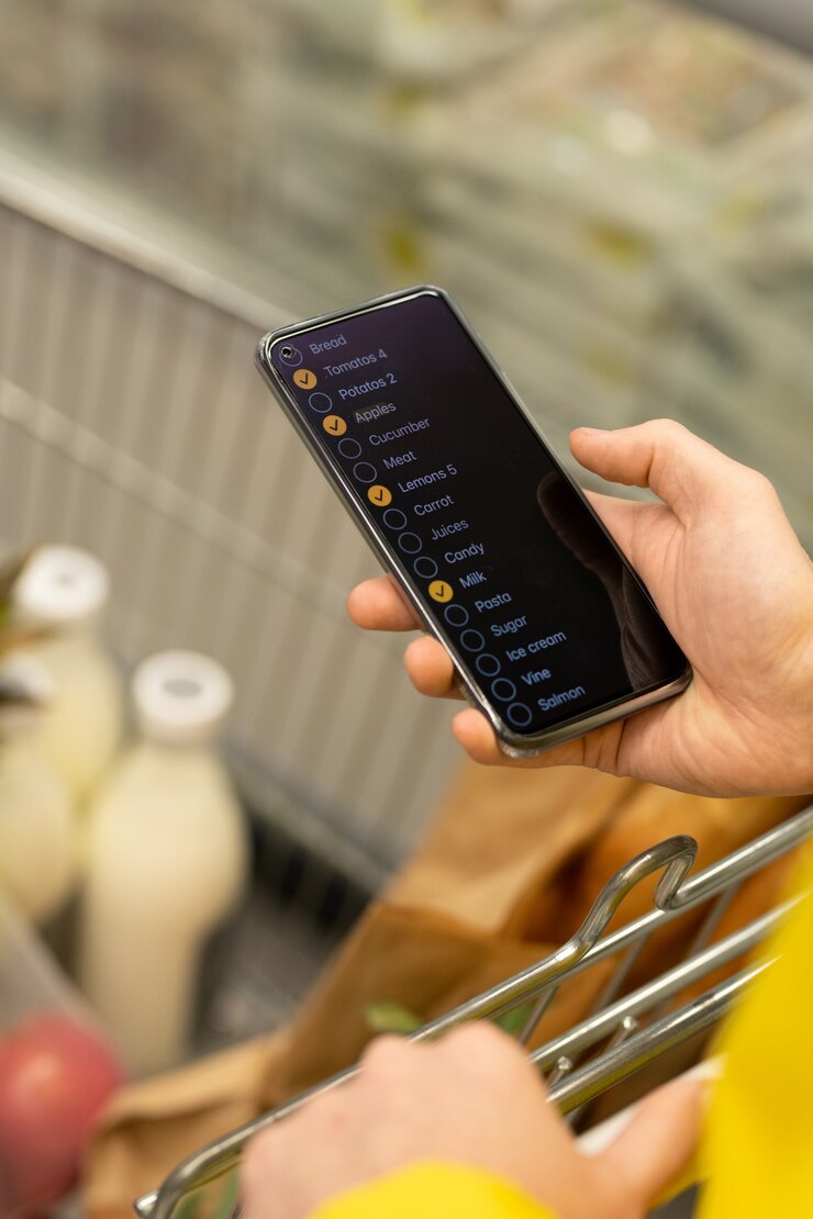 Foto mensajero de sexo masculino joven con smartphone desplazándose a través de la lista de cosas para comprar en carrito de compras con pan fresco y leche en el supermercado