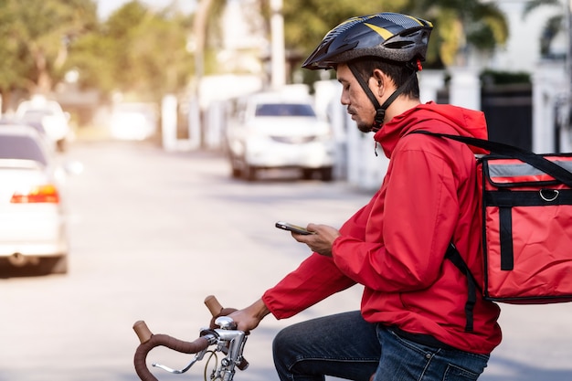 Mensajero en uniforme rojo con una caja de entrega en la espalda montando una bicicleta y mirando en el teléfono celular para verificar la dirección para entregar la comida al cliente. Mensajero en bicicleta entregando comida en la ciudad.