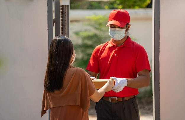 Un mensajero con uniforme rojo con máscara protectora y guantes lleva el paquete a casa del cliente.