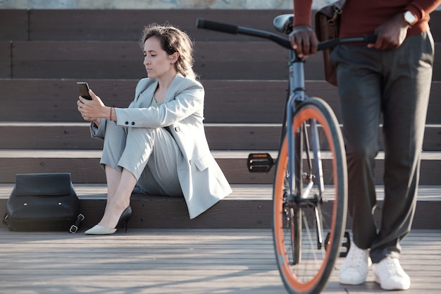 Mensajes de texto de la empresaria seria en el teléfono inteligente con el hombre empujando la bicicleta en la parte delantera