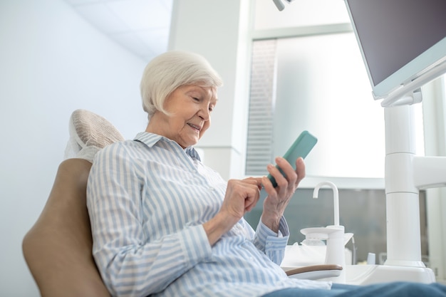 Mensajes de texto. Mujer mayor sentada en la oficina de dentistas y usando su teléfono inteligente