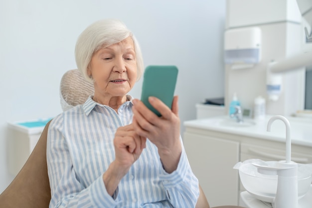 Mensajes de texto. Mujer mayor sentada en la oficina de dentistas y usando su teléfono inteligente