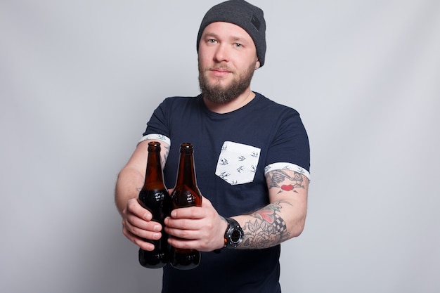 Menschen, 4K und Lifestyle-Konzept - Brutaler bärtiger Mann mit tätowiertem Arm trinkt ein Bier aus einer Flasche. gut aussehender Mann mit Hut. Brutaler bärtiger Junge mit Tattoo