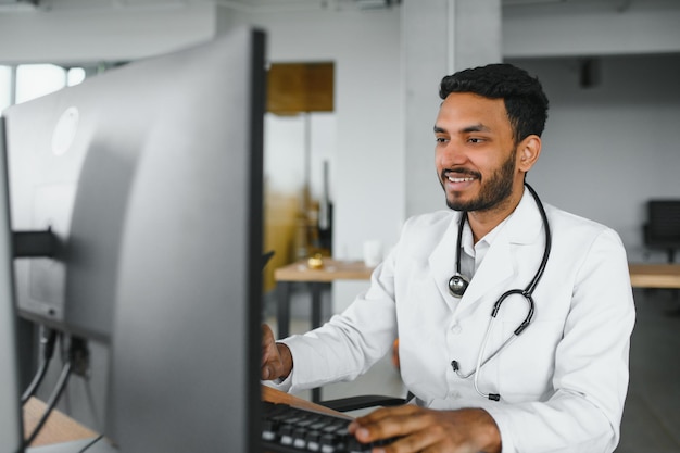 Menschen Beruf und Medizin Konzept Lächelnder männlicher indisch-arabischer Arzt im weißen Mantel, der in der Arztpraxis am Schreibtisch mit Laptop sitzt