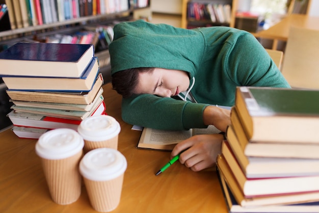 menschen, bildung, sitzung, prüfungen und schulkonzept - müder schüler oder junger mann mit büchern und kaffee, der in der bibliothek schläft